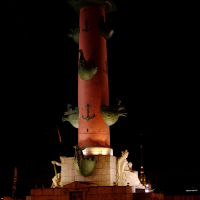 Illuminated rostral column in St. Petersburg, Russia, on a clear night, showcasing its historic sculptures and the beacon light on top, against the backdrop of a dark sky.