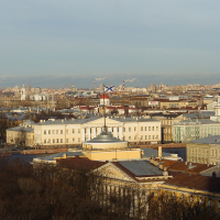 A panoramic view of St. Petersburg's cityscape with historic buildings under a soft sky, industrial cranes in the distance, and the foreground featuring autumnal trees, suggesting a blend of urban life and