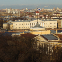 An aerial view of a historic neoclassical building in St. Petersburg basking in the golden hues of the setting sun, with a sprawling cityscape and cloudy skies in the backdrop.