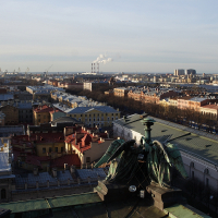 A panoramic view of St. Petersburg with historic architecture, featuring rooftops under a clear sky, with industrial smokestacks in the distance, as seen from an elevated position.