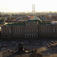 Aerial view of a historical square in St. Petersburg at sunset, featuring a grand building with a green rooftop and flag, a prominent statue, and neatly parked cars, against a cityscape backdrop with