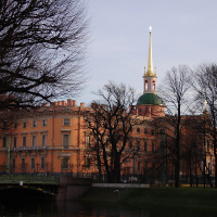 Historic orange building with a green dome and golden spire stands by a serene water canal in St. Petersburg, with a bridge in the foreground amidst leafless trees under a soft dusk sky.