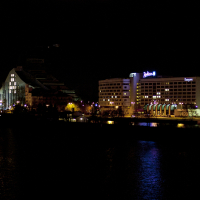 Nighttime image of a cityscape featuring two prominent, illuminated buildings by a body of water. On the left, the building has a modern, triangular design. On the right, a hotel with visible signage. Reflections of the lights shimmer on the dark water's surface during Staro Rīga 2011 festivities.