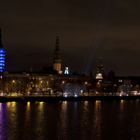 A nighttime cityscape featuring a skyline with various illuminated towers and spires showcases the charm of Staro Rīga 2011. A large building on the left glows blue, casting reflections on a river in the foreground. The dark sky is dotted with scattered clouds, while streetlights line the riverside, illuminating the scene below.