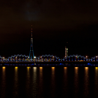 A nighttime view of the city skyline during the 2011 Staro Rīga festival features a brightly lit bridge spanning a river, with arched sections illuminated in blue and yellow lights. A tall tower with red lights at the top stands prominently in the background, and city buildings with lights on are visible to the right.
