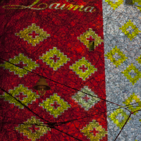 A brightly illuminated wall at the 2011 Staro Rīga festival displays a colorful pattern with red and yellow diamond shapes and an ornate script reading “Laima” at the top. Part of the wall is covered in brickwork, while the other part has a grayish texture. Vines and light fixtures are visible against the lit background.