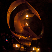 A metal sail-like sculpture illuminated by small lanterns is pictured at night. The abstract structure features curved metal sheets forming a spiral with glowing flames inside. In the background, silhouettes of people holding colorful lights are visible against a dark, blurry forest backdrop at Staro Rīga 2011.