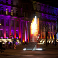 A nighttime scene from 2011 features a tall building illuminated by vibrant pink and purple lights. In front of the building is a modern art installation with vertical, illuminated rods in varying heights, resembling flames. The surrounding area is busy with people and vehicles, and colorful street signs are visible. This scene is part of the Staro Rīga festival.