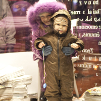 Two young children stand inside a brightly lit space with large windows. The older child, wearing a purple hooded coat, smiles and stands behind a smaller child in a brown snowsuit holding a toy on a string. Stacks of newspapers lie on the floor nearby. Snowflakes from the 2011 Staro Rīga festival can be seen falling outside.