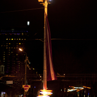 A streetlight illuminates the dark street with a warm, amber glow. A multicolored flag hangs from the pole. Below, traffic trails captured in long exposure appear as bright, colorful streaks. In the background, buildings and additional lights are visible against the night sky of Staro Rīga 2011.