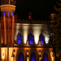A nighttime image of a historic Gothic-style building with arched windows illuminated by blue and white lights during Staro Rīga 2011. A large pole with a red and white banner is in the foreground. Several people in winter clothing are gathered near the building, and the scene is adorned with a nearby tree.