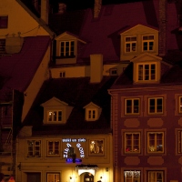 A nighttime street scene shows people walking past a row of charming, old-fashioned buildings with lit windows. One building features a brightly illuminated sign in blue with white text. The atmosphere, reminiscent of Staro Rīga 2011, is warm and inviting, with light spilling onto the cobblestone street.