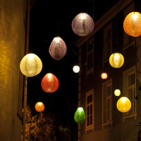 Colorful spherical lanterns in shades of yellow, purple, red, green, and orange hang above a narrow street at night during the 2011 Staro Rīga festival. The lanterns illuminate the area with a soft, warm glow against the backdrop of dark buildings. The scene evokes a festive and cozy ambiance.