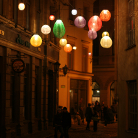 A warmly lit narrow street at night, featuring hanging colorful lanterns above. People walk on the cobblestone pathway lined with old buildings and shops. The atmosphere is cozy and inviting, with the soft glow of lanterns illuminating the scene—a snapshot reminiscent of Staro Rīga in 2011.