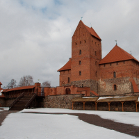 A medieval brick castle in Trakai, Lithuania, with distinctive red-tiled roofs stands against a cloudy winter sky, with remnants of snow covering the ground, suggesting a chilly and quiet historical ambiance.