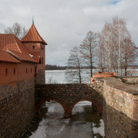 A medieval Trakai Castle with terracotta roofs stands beside a tranquil lake in Lithuania, its ancient stonework bridge dusted with snow, beneath a cloudy winter sky.