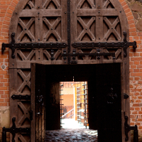 An imposing medieval wooden gate of Trakai Castle stands ajar, revealing a glimpse of a sunlit courtyard beyond, contrasting the shadowed, time-worn entrance with its iron reinforcements and studded