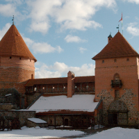 Medieval Trakai Castle in winter with snow-covered ground, featuring distinctive red-brick towers and walls under a partly cloudy sky, embodying historical architecture in Lithuania.