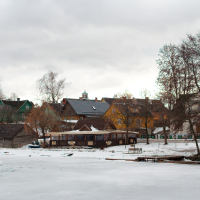 A winter scene of a quaint village with snow-covered roofs, the frozen river forefront near Trakai Castle, bare trees, and a cloudy sky suggesting a serene, chilly day in Lithuania.