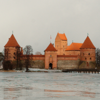 A medieval castle, known as Trakai, with distinctive red-orange walls and pointed towers stands majestically amidst a wintry landscape, its reflection blurred on the icy surface of a surrounding moat