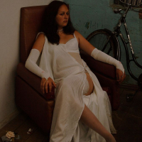 A woman in a white dress and long gloves sits on a brown chair in a dimly lit room. She appears thoughtful and relaxed, with one leg crossed over the other. Behind her, a bicycle is partially visible, leaning against a teal wall adorned with upuris mākslu. The floor has scattered items, including bottles and paper.
