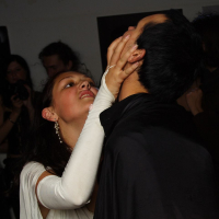 A woman wearing a white outfit and long white gloves is holding the face of a man dressed in black. Her expression is intense, her fingers pressing into his cheeks as she leans towards him, capturing a moment of raw emotion akin to māksla. The background is dimly lit with a few indistinct people visible.