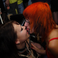 Two individuals with brightly colored hair are engaging in an intimate moment. One has black hair, the other has orange. They lean close, tongues out towards each other. The dimly lit background, peppered with blurry figures, suggests a social setting where Upuris prasa mākslu is the unspoken theme.