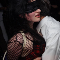 A blindfolded person with long dark hair wearing a fishnet top and a red corset is entwined with ropes and embraces another person. The person being held appears calm. Several other people are visible in the background, creating a party or gathering atmosphere, embodying the spirit of Upuris prasa mākslu.
