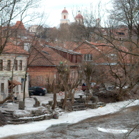 A tranquil, snowy scene in Vilnius with bare trees, a meandering stream, and traditional buildings, with a church's domes peeking in the distance.