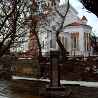 A statue stands in the foreground of a serene winter scene, with a historic church nestled among bare branches in the background, under a cloudy sky in Vilnius.