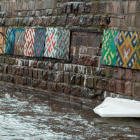 Colorful knitwear pieces displayed on an old brick wall beside a chilly river in Vilnius, showcasing a harmonious blend of art and nature with remnants of snow on the bank.