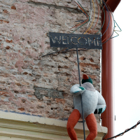 A whimsical welcome sign featuring a stylized metallic representation of a bird wearing a hat perched atop, with colorful wire accents, positioned against a weathered wall in Vilnius with exposed brick textures