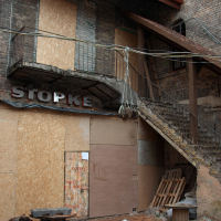 An old, dilapidated building in Vilnius with a broken wooden staircase and boarded-up windows, featuring graffiti and a faded "stopke" sign, suggesting a forgotten or neglected urban space.