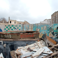 A weathered boat rests amidst urban decay and debris in Vilnius, with graffiti art on a neglected building providing a bleak yet colorful backdrop in a desolate setting.