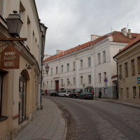 A cobbled street winds through the old town of Vilnius, showcasing historic architecture with muted tones. Pedestrians stroll past quaint buildings, while cars line the edges of the relaxed urban scene.