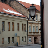 An old-fashioned street lamp emerges from a wall, overlooking a quiet, cobblestoned street with traditional Vilnius buildings, under a soft evening sky.