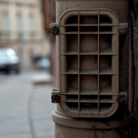A weathered and dusty exhaust vent with a protective metal grid is attached to a brick wall on an urban street in Vilnius, with blurred vehicles visible in the background, suggesting a bustling city atmosphere