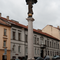 An angel statue with outstretched wings stands atop a column on a cobblestone street in Vilnius, flanked by classical European architecture under a cloudy sky.