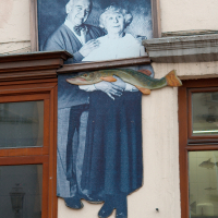 A vintage black-and-white photo of a smiling couple is displayed above a window in Vilnius, with a three-dimensional fish-themed ornament beneath it, creating the quirky illusion of the couple holding the fish
