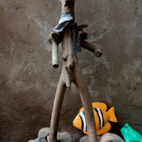 A whimsical wooden sculpture donning a cap and scarf stands amidst stones alongside a colorful fish toy and a green frog in Vilnius, invoking a playful garden scene with a touch of rustic charm.