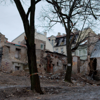 A somber urban scene in Vilnius showing a demolished building amidst surviving structures, with exposed rubble and leafless trees under an overcast sky, evidencing urban decay or recent reconstruction efforts.