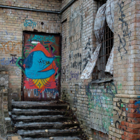 A weathered urban staircase in Vilnius, Lithuania, leads to a graffiti-tagged door, surrounded by crumbling brick walls and a tattered metal awning, evoking a scene of decay and