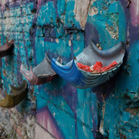 A weathered urban wall in Vilnius displaying a creative public art installation, featuring a series of half pipes painted in various colors, repurposed as planters against a backdrop of faded blue and