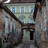 A narrow alleyway between weathered walls adorned with graffiti in Vilnius leads to a green three-story building with an archway entrance, showcasing a blend of urban decay and historical architecture.