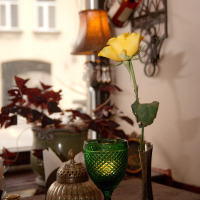 A single yellow rose stands in a slender metallic vase beside a green glass goblet and decorative items on a table, with a softly lit lamp and a violin in the blurred background creating a warm, inviting