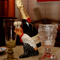 A whimsical figurine of a chef kissing a bottle of moët champagne, surrounded by elegant glassware and tiny decorative details on a warm, intimate tabletop setting in Vilnius.