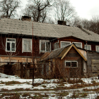 A dilapidated wooden house with peeling paint and a sagging roof stands in disrepair amid a sparse snowy landscape in Vilnius, surrounded by a wire fence, evoking a sense of