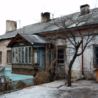 A weathered courtyard in Vilnius with a dilapidated building, featuring a wooden extension and a lone bare tree. A small, colorful doghouse adds a touch of warmth to the otherwise bleak