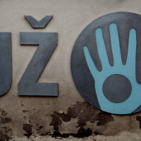 An aged wall in Vilnius features the letters "uz" next to a circular metal sign with a stylized white handprint, suggesting a logo or symbol, all under a weathered "y