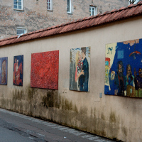 An outdoor urban art gallery in Vilnius featuring a row of diverse paintings hung on a weathered wall, adding a splash of cultural vibrancy to a muted street.